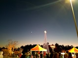 An evening scene featuring a festive gathering with people under brightly colored tents. A tall, illuminated structure resembling a Christmas tree with multicolored lights stands in the background, with a large menorah lit nearby. The sky transitions from deep blue to a lighter gradient near the horizon, and a crescent moon is visible.