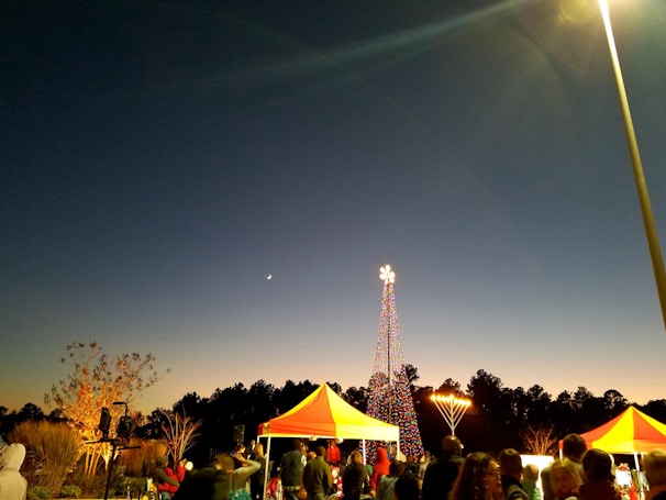 An evening scene featuring a festive gathering with people under brightly colored tents. A tall, illuminated structure resembling a Christmas tree with multicolored lights stands in the background, with a large menorah lit nearby. The sky transitions from deep blue to a lighter gradient near the horizon, and a crescent moon is visible.