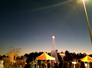An evening scene featuring a festive gathering with people under brightly colored tents. A tall, illuminated structure resembling a Christmas tree with multicolored lights stands in the background, with a large menorah lit nearby. The sky transitions from deep blue to a lighter gradient near the horizon, and a crescent moon is visible.