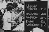 A group of people are gathered at a market stall examining packaged goods. Next to them is a chalkboard menu listing items such as crepes and fruit salad with their respective prices in what appears to be an Indonesian currency format.
