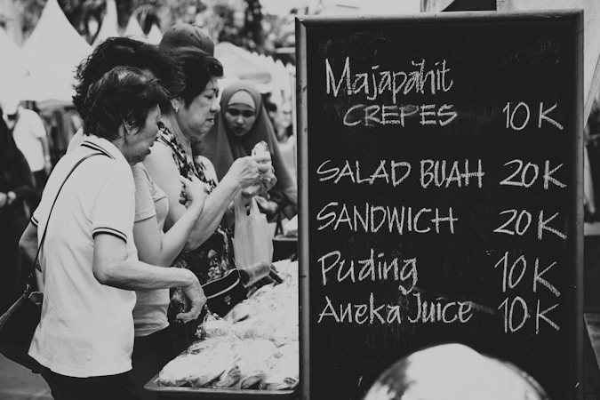 A group of people are gathered at a market stall examining packaged goods. Next to them is a chalkboard menu listing items such as crepes and fruit salad with their respective prices in what appears to be an Indonesian currency format.