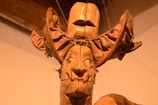 A wooden sculpture consists of intricately carved features with an exaggerated headpiece. The artwork appears to have a mask-like face with distinct facial features, possibly influenced by tribal or ethnic art styles. The material showcases rich textures and a warm, earthy tone.