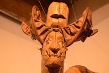 A wooden sculpture consists of intricately carved features with an exaggerated headpiece. The artwork appears to have a mask-like face with distinct facial features, possibly influenced by tribal or ethnic art styles. The material showcases rich textures and a warm, earthy tone.