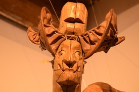 A wooden sculpture consists of intricately carved features with an exaggerated headpiece. The artwork appears to have a mask-like face with distinct facial features, possibly influenced by tribal or ethnic art styles. The material showcases rich textures and a warm, earthy tone.