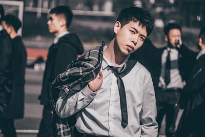 A young man stands with a casual yet stylish pose, holding a plaid coat over his shoulder. He wears a button-up shirt with a tie and gazes directly at the viewer. Several other people are in the background, blurred and walking by.