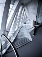An architectural interior with angular geometric designs featuring a staircase flanked by metal railings. The space is defined by the convergence of modern materials like glass and concrete, highlighted by the interplay of light and shadows.