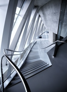 An architectural interior with angular geometric designs featuring a staircase flanked by metal railings. The space is defined by the convergence of modern materials like glass and concrete, highlighted by the interplay of light and shadows.