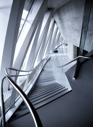 An architectural interior with angular geometric designs featuring a staircase flanked by metal railings. The space is defined by the convergence of modern materials like glass and concrete, highlighted by the interplay of light and shadows.