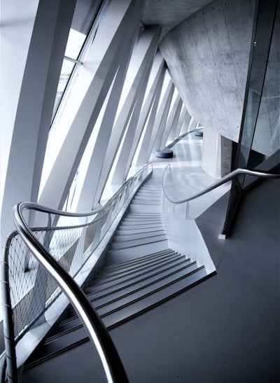 An architectural interior with angular geometric designs featuring a staircase flanked by metal railings. The space is defined by the convergence of modern materials like glass and concrete, highlighted by the interplay of light and shadows.