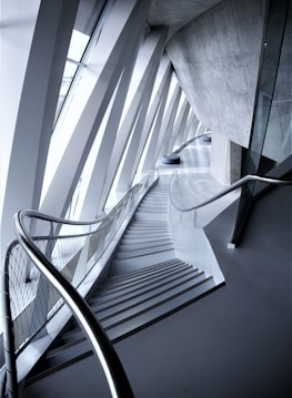 An architectural interior with angular geometric designs featuring a staircase flanked by metal railings. The space is defined by the convergence of modern materials like glass and concrete, highlighted by the interplay of light and shadows.