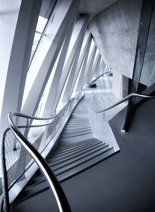 An architectural interior with angular geometric designs featuring a staircase flanked by metal railings. The space is defined by the convergence of modern materials like glass and concrete, highlighted by the interplay of light and shadows.