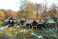 A series of small wooden huts is perched over a stream with cascading waterfalls, set against a backdrop of autumnal trees with colorful foliage. People are walking around, adding a sense of life to the picturesque natural setting.