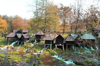 A series of small wooden huts is perched over a stream with cascading waterfalls, set against a backdrop of autumnal trees with colorful foliage. People are walking around, adding a sense of life to the picturesque natural setting.