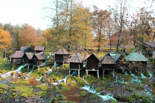 A series of small wooden huts is perched over a stream with cascading waterfalls, set against a backdrop of autumnal trees with colorful foliage. People are walking around, adding a sense of life to the picturesque natural setting.
