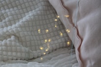 A cozy setting featuring a white textured blanket and a light pink pillow. Warm yellow lights are softly glowing on the blanket, creating a calming and inviting atmosphere.