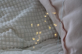 A cozy setting featuring a white textured blanket and a light pink pillow. Warm yellow lights are softly glowing on the blanket, creating a calming and inviting atmosphere.