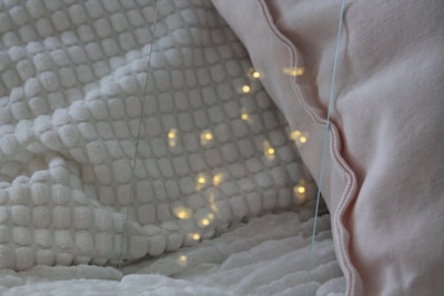 A cozy setting featuring a white textured blanket and a light pink pillow. Warm yellow lights are softly glowing on the blanket, creating a calming and inviting atmosphere.