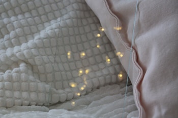 A cozy setting featuring a white textured blanket and a light pink pillow. Warm yellow lights are softly glowing on the blanket, creating a calming and inviting atmosphere.