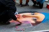 A street artist is creating a detailed chalk drawing on pavement resembling a famous painting. The drawing features a female face with a blue headscarf and expressive eyes. Several pieces of colored chalk are scattered nearby. Other people are gathered around, observing the artwork.