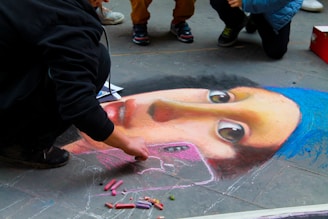 A street artist is creating a detailed chalk drawing on pavement resembling a famous painting. The drawing features a female face with a blue headscarf and expressive eyes. Several pieces of colored chalk are scattered nearby. Other people are gathered around, observing the artwork.