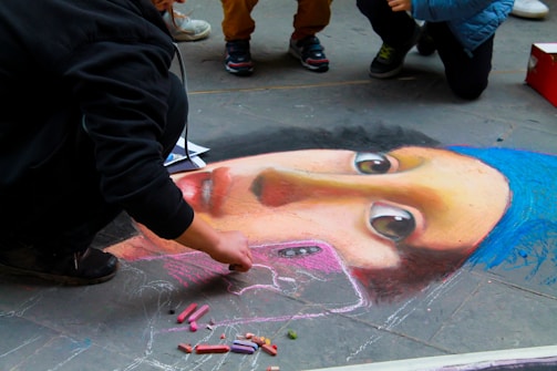 A street artist is creating a detailed chalk drawing on pavement resembling a famous painting. The drawing features a female face with a blue headscarf and expressive eyes. Several pieces of colored chalk are scattered nearby. Other people are gathered around, observing the artwork.