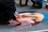 A street artist is creating a detailed chalk drawing on pavement resembling a famous painting. The drawing features a female face with a blue headscarf and expressive eyes. Several pieces of colored chalk are scattered nearby. Other people are gathered around, observing the artwork.