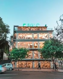 A multi-story building with large glass windows reveals a showroom filled with a variety of lights and chandeliers. The structure is illuminated warmly, and a bright green sign 'EGLO' is displayed at the top. The street in front features trees and moving vehicles, with a modern urban atmosphere.