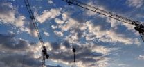 Several large construction cranes are silhouetted against a backdrop of a cloudy sky, with a mix of light and dark clouds scattered across the blue expanse.
