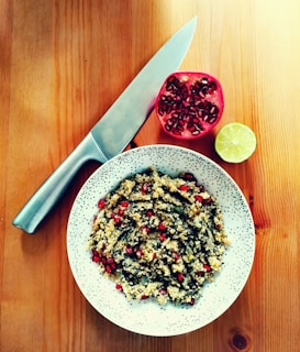 A bowl of couscous mixed with herbs and pomegranate seeds is placed on a wooden table. Next to it, half a pomegranate and half a lime are visible. A large chef’s knife lies nearby, showcasing the preparation area for a delicious meal.
