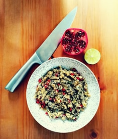Un bol de couscous mélangé avec des herbes et des graines de grenade est posé sur une table en bois. À côté, on voit une moitié de grenade et une moitié de citron vert. Un grand couteau de chef est posé à proximité, illustrant l'espace de préparation pour un délicieux repas.