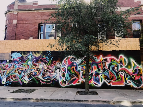A vibrant graffiti mural covers the wall of a brick building. The artwork features an array of intricate shapes, arrows, and abstract forms in bright colors such as blue, purple, green, and pink. A tree stands in front of the mural, partially obscuring the view, with its branches and leaves casting shadows on the artwork.