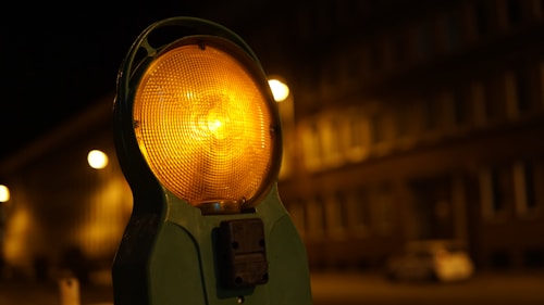 A close-up of an orange construction warning light illuminated at night, casting a warm glow. In the background, there is a blurred view of a building with multiple windows, softly lit by street lights, creating a nighttime urban setting.