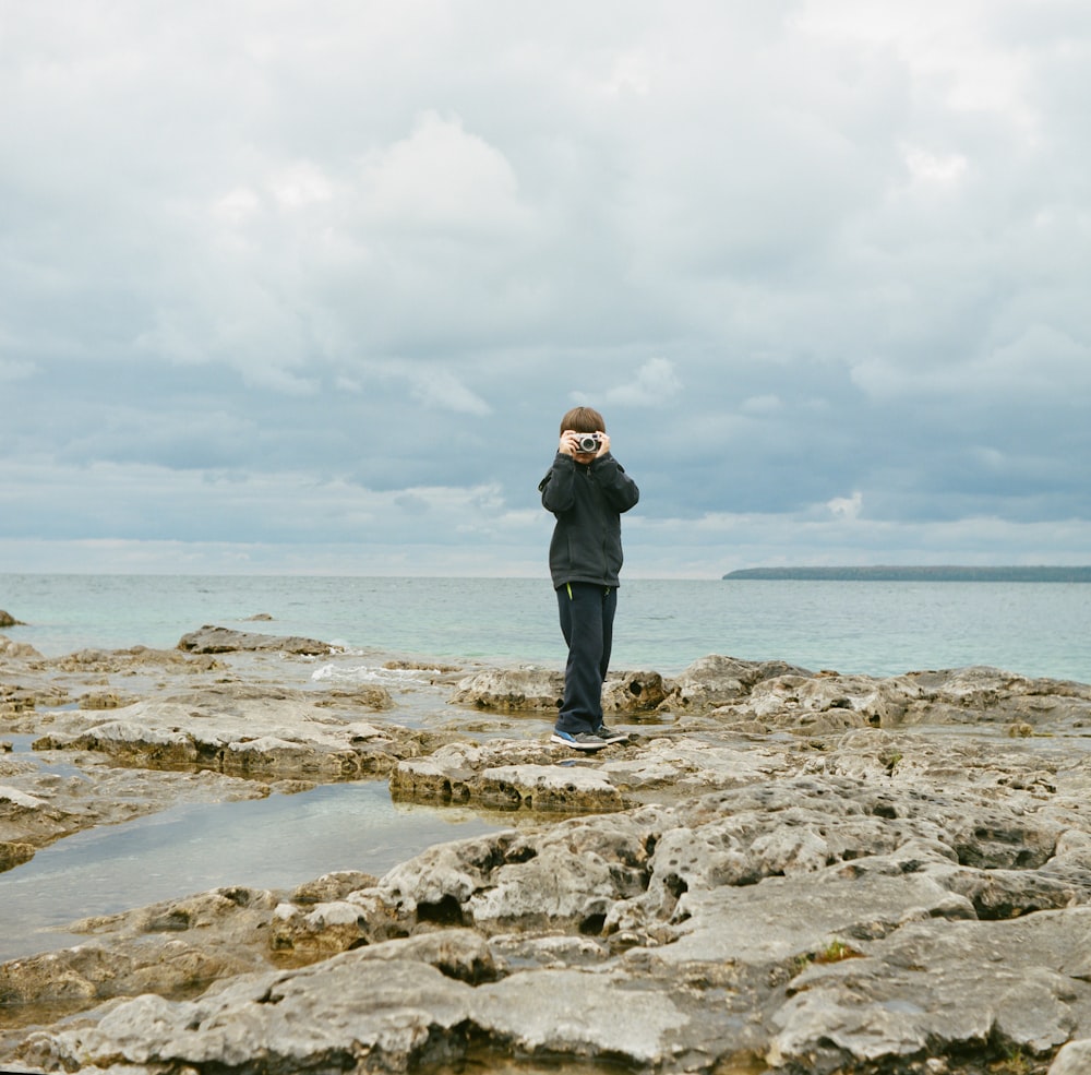 man standing on rock formation near body of water holding camera