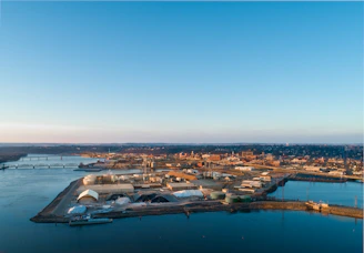 An industrial area situated along a waterfront with several buildings and storage facilities. A bridge spans across the water in the background, and a cityscape is visible in the distance. The sky is clear with a hint of sunset, casting warm light over the scene.