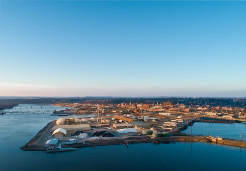 An industrial area situated along a waterfront with several buildings and storage facilities. A bridge spans across the water in the background, and a cityscape is visible in the distance. The sky is clear with a hint of sunset, casting warm light over the scene.