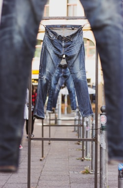 Several pairs of blue jeans are hung on metal racks outdoors. The background shows a building with arched windows and other elements of an urban environment. The jeans appear to be placed in a line, creating a visual pathway through the racks.