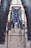Several pairs of blue jeans are hung on metal racks outdoors. The background shows a building with arched windows and other elements of an urban environment. The jeans appear to be placed in a line, creating a visual pathway through the racks.
