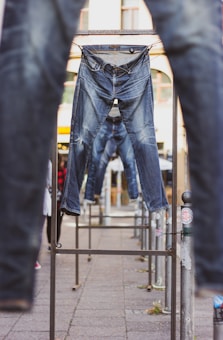 Several pairs of blue jeans are hung on metal racks outdoors. The background shows a building with arched windows and other elements of an urban environment. The jeans appear to be placed in a line, creating a visual pathway through the racks.
