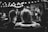 A black and white photo of a couple sitting closely on a couch, facing away. Soft, blurred lights hang above, creating a romantic atmosphere. The focus is on the back of their heads, with blurred figures in the background suggesting a gathering or event.