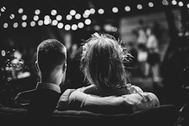 A black and white photo of a couple sitting closely on a couch, facing away. Soft, blurred lights hang above, creating a romantic atmosphere. The focus is on the back of their heads, with blurred figures in the background suggesting a gathering or event.