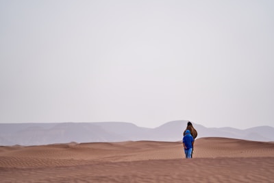 A person dressed in blue walks alongside a camel across expansive sand dunes, with gentle rolling hills visible in the distant background beneath a pale sky.