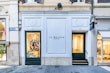 A storefront with a white facade and black signage displaying 'Jo Malone London' between two numbered doors 42 and 43. The left side features a window display with colorful decorations and a smiling person visible inside. Adjacent to this, another store window shows more neutral colors. The street is paved and clean, with nearby structures appearing elegant and well-maintained.