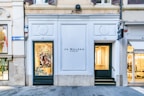 A storefront with a white facade and black signage displaying 'Jo Malone London' between two numbered doors 42 and 43. The left side features a window display with colorful decorations and a smiling person visible inside. Adjacent to this, another store window shows more neutral colors. The street is paved and clean, with nearby structures appearing elegant and well-maintained.