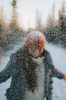 A person in winter clothing stands amidst a flurry of snow, with trees blurred in the background. The motion blur suggests movement, and the focus on the snow creates a dynamic and energetic scene.