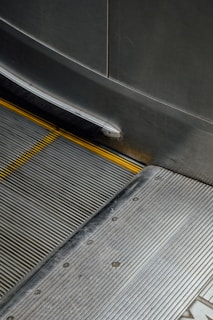 An image of an escalator with metal steps and yellow safety stripes along the edges. The surrounding paneling is also metallic, reflecting a slightly industrial look.