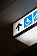 A directional overhead sign indicating an accessible path is visible, featuring a black arrow pointing upwards on a white background. The sign includes the universal symbol for accessibility with a person in a wheelchair, depicted in white against a blue square.