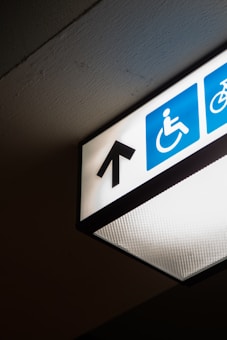 A directional overhead sign indicating an accessible path is visible, featuring a black arrow pointing upwards on a white background. The sign includes the universal symbol for accessibility with a person in a wheelchair, depicted in white against a blue square.