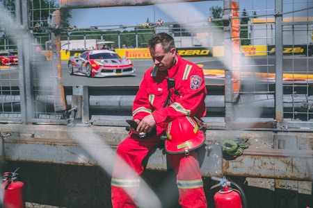A person wearing red firefighter gear is sitting on a concrete barrier near a racetrack. Several race cars are visible in the background on the track. The person appears to be using a smartphone and has a red helmet placed on their lap. Fire extinguishers are placed nearby.