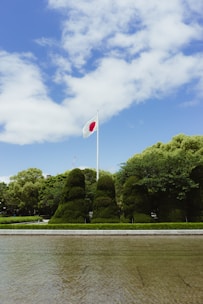 A flagpole with the Japanese flag waves against a backdrop of a clear blue sky and scattered white clouds. Below, manicured green bushes and trees are neatly arranged, with a paved path and a body of water in the foreground.