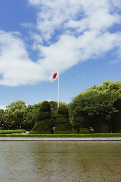 A flagpole with the Japanese flag waves against a backdrop of a clear blue sky and scattered white clouds. Below, manicured green bushes and trees are neatly arranged, with a paved path and a body of water in the foreground.